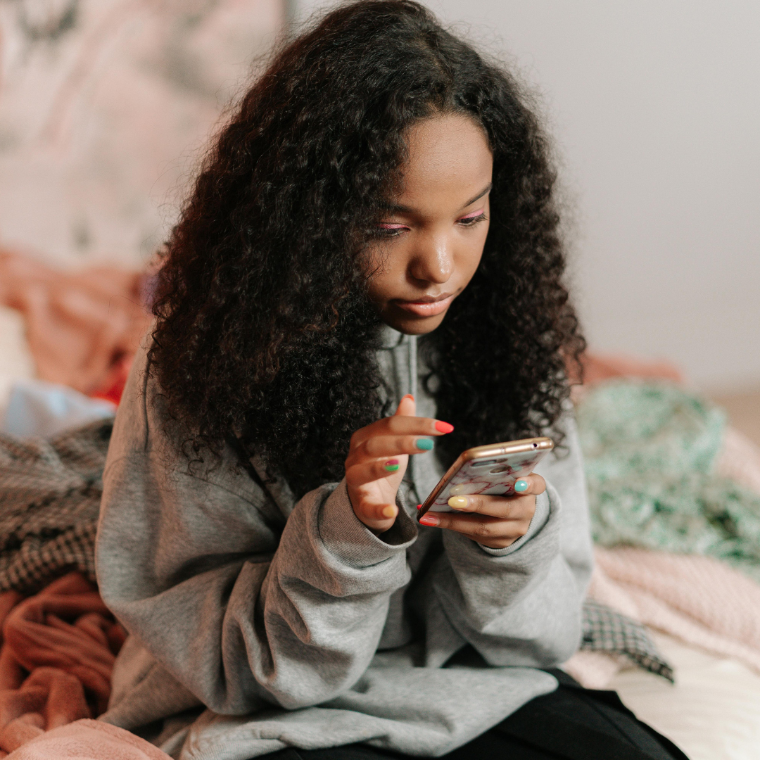 Teenage girl with curly hair sitting on a bed, looking closely at her phone, wearing a grey sweatshirt with colourful nail polish.