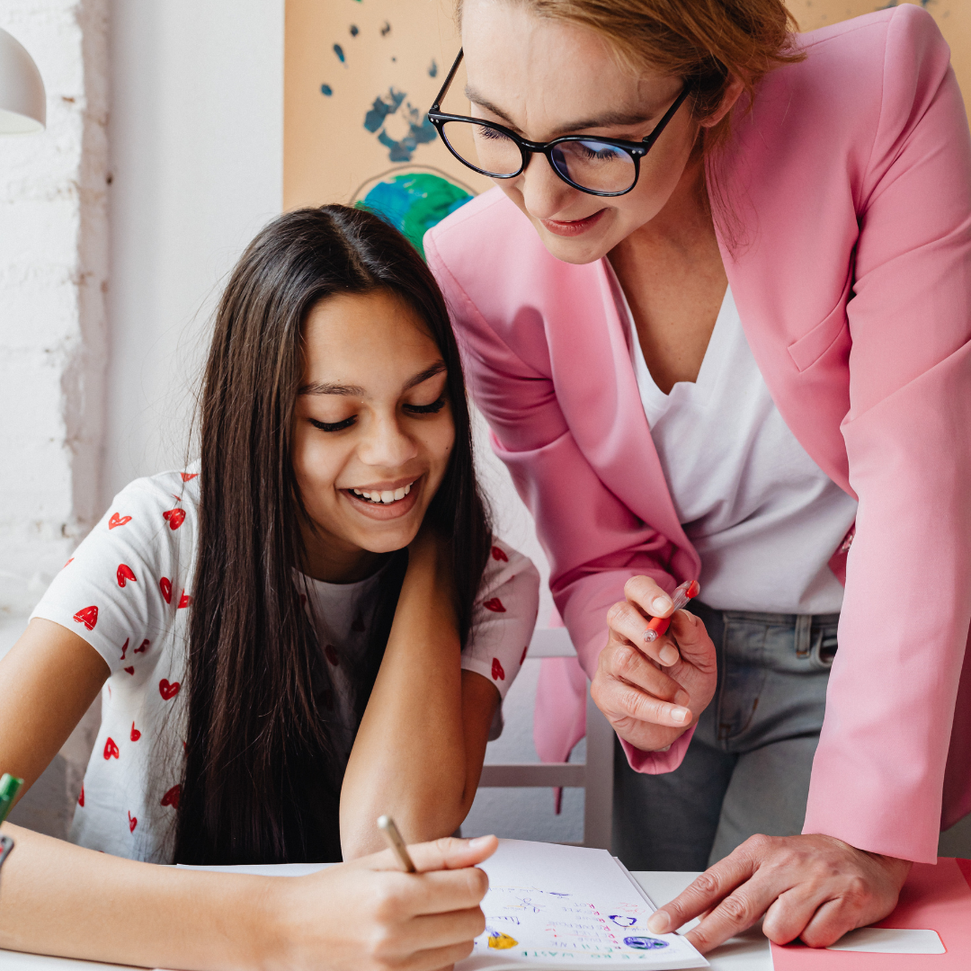 Teacher in a pink blazer leaning over to help a smiling girl with her writing at a desk.