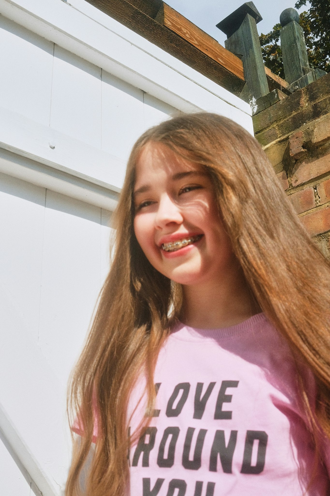 Smiling teenage girl with braces and long brown hair, wearing a pink "Love around you" T-shirt outdoors.