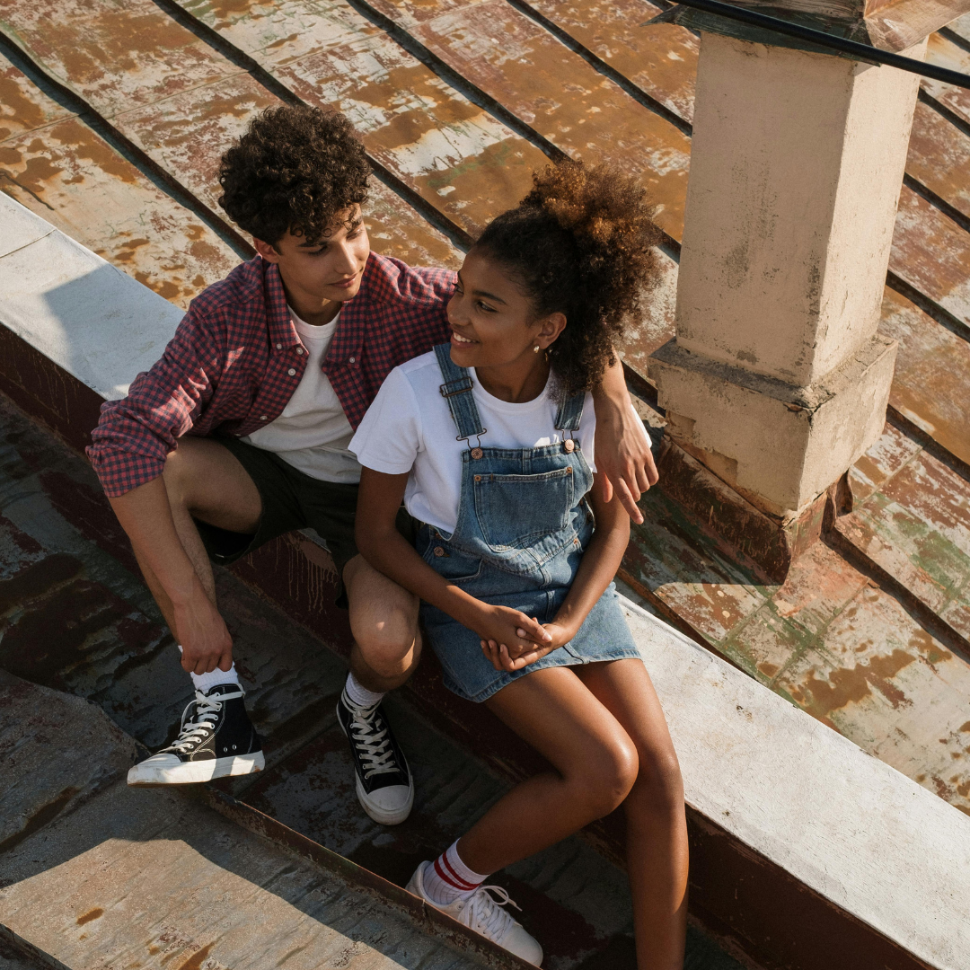 Teenage couple sitting close together on a rooftop, the boy with his arm around the girl, both smiling at each other.