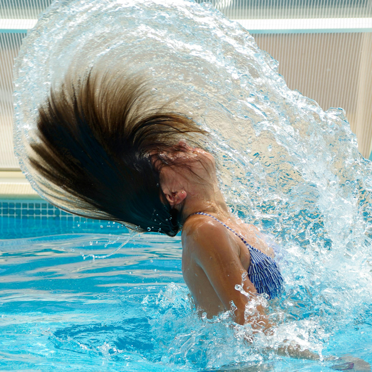 Girl in a pool flipping her wet hair back, sending up a large splash of water.