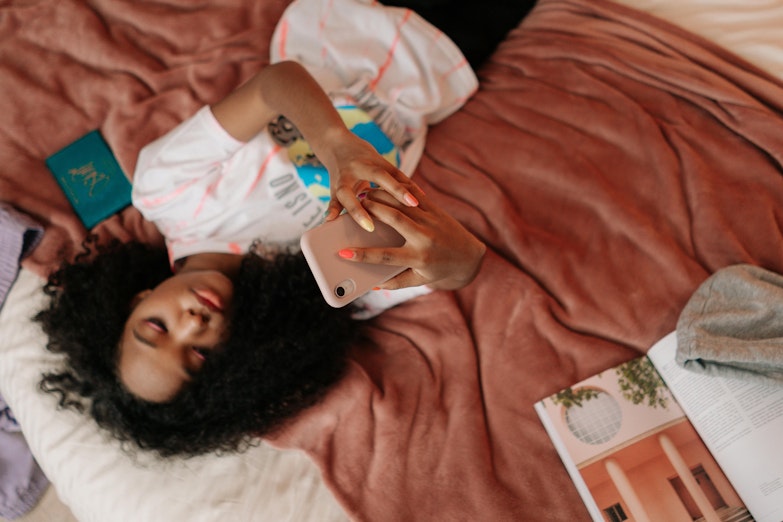 Girl with curly hair lying on a bed, taking a photo of herself on her phone.
