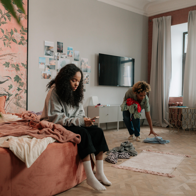 Teenage girl sitting on her bed looking at her phone while her mother picks up clothes from a messy bedroom floor.