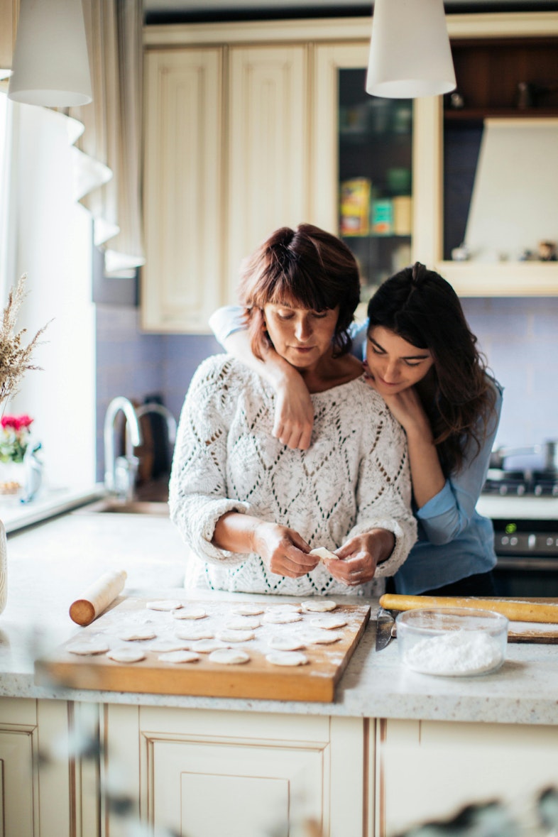 Teenage girl hugging her mother from behind while she cuts dough into circles at a kitchen counter.