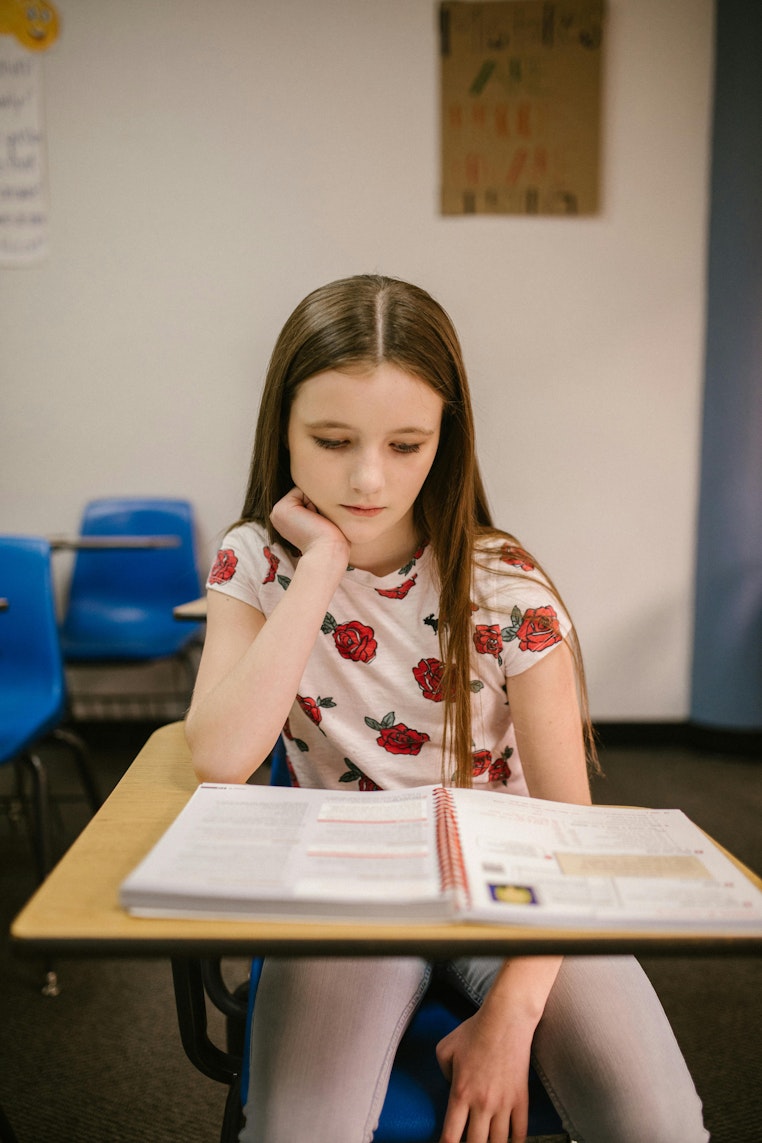 Girl resting her head on her hand at a classroom desk, looking down at an open workbook.