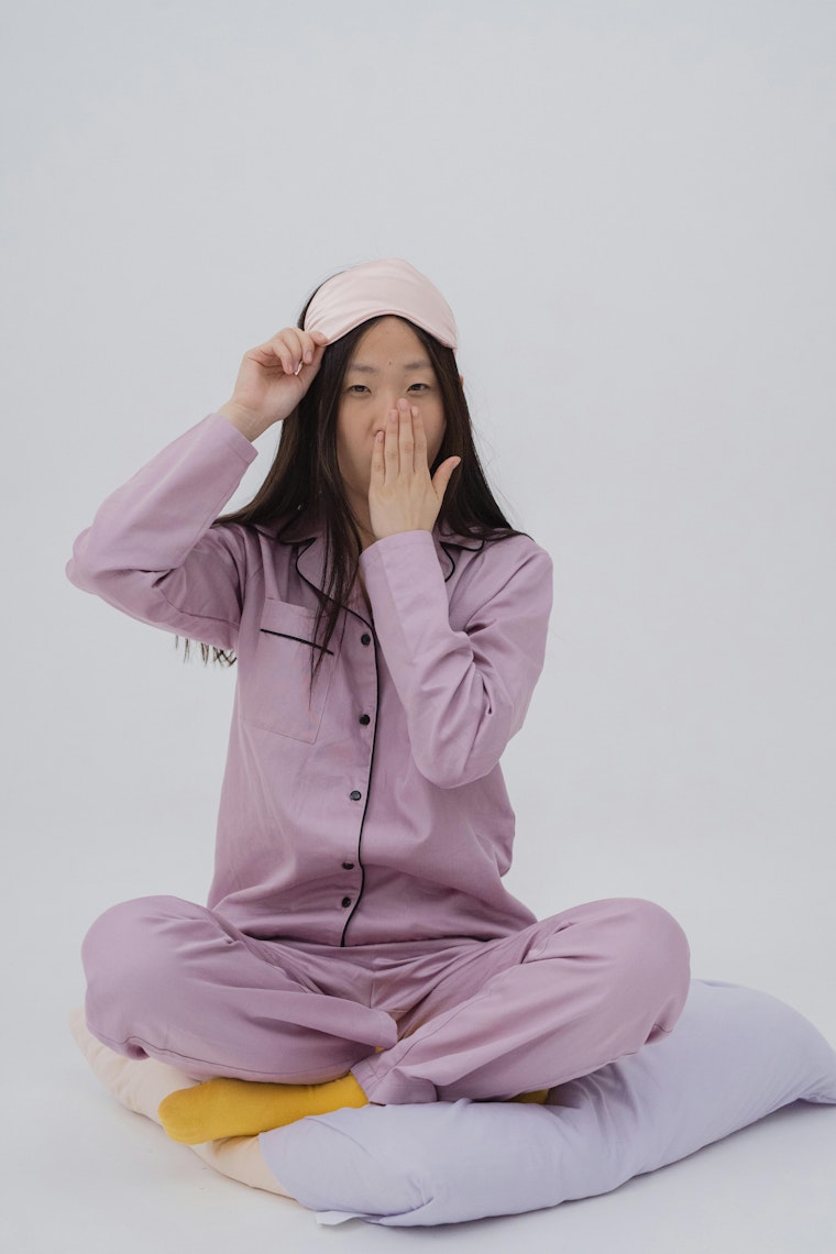 Teenage girl in purple pyjamas sitting cross-legged, lifting a sleep eye mask off her forehead and covering her mouth with her hand.