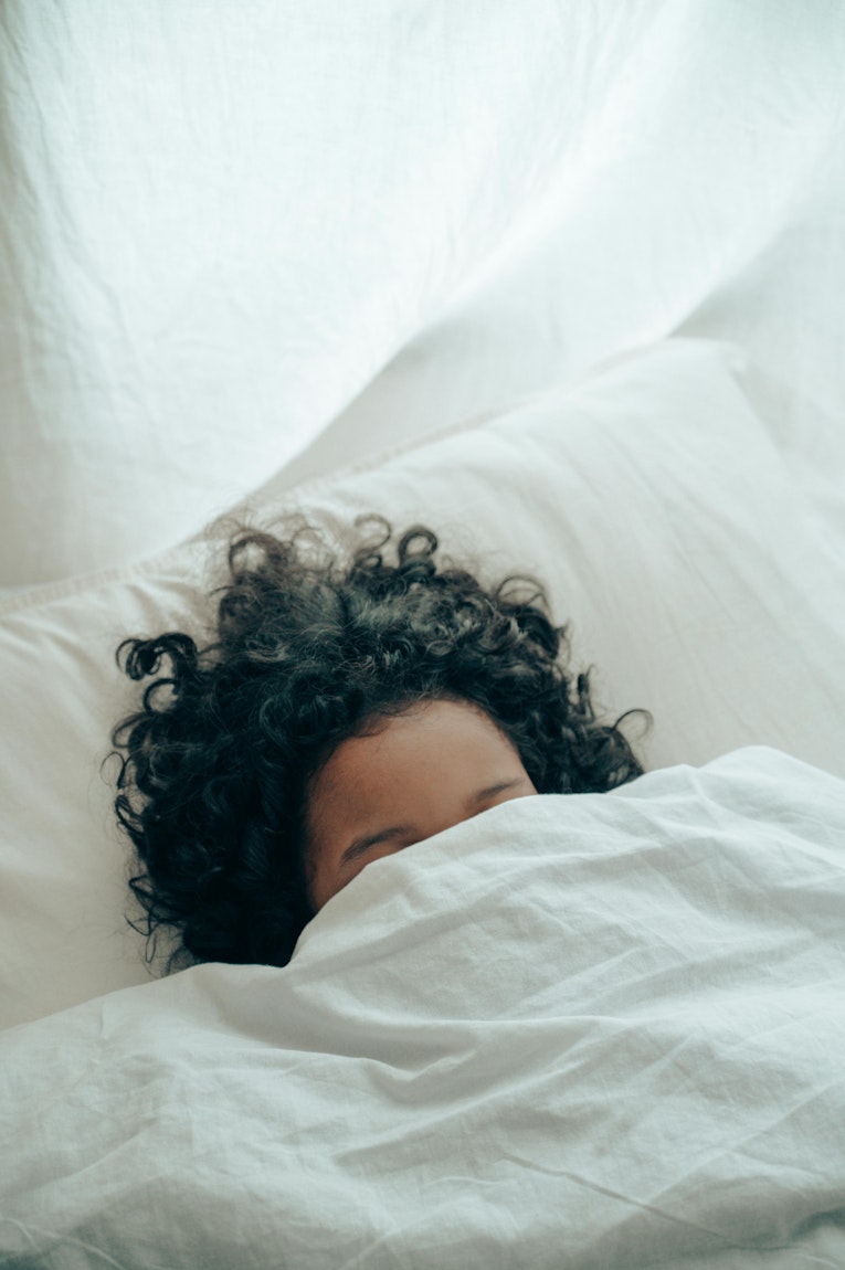 Overhead view of a teenager with curly hair tucked under a white duvet, with only the top of their head visible.