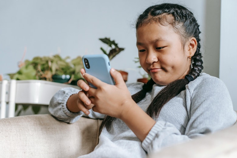 Young girl with braided hair sitting on a sofa, pulling a face while scrolling on her phone, with plants in the background.