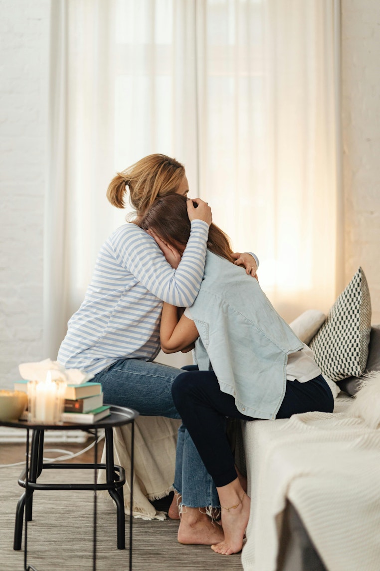 Mother sitting on a bed, hugging her daughter who has her head resting against her chest, in a cosy candlelit room.