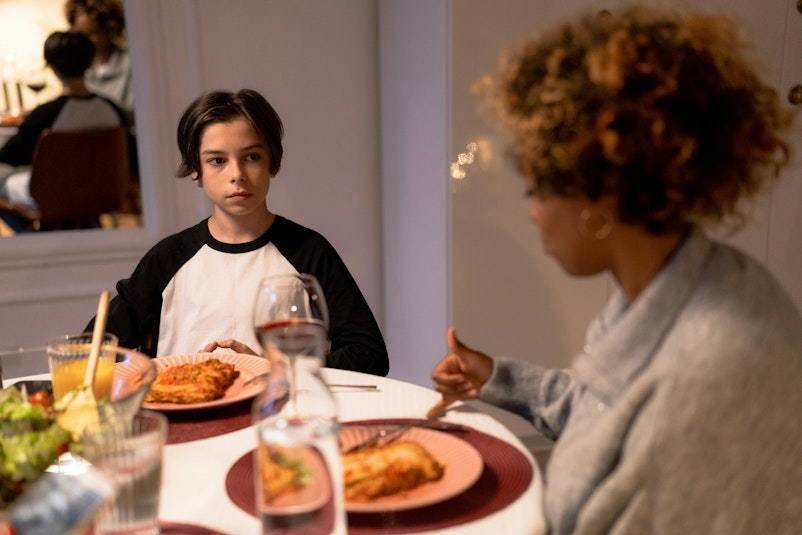 Woman with curly hair talking with her hands to her daughter's boyfriend, sitting across the dinner table, who looks at her with a serious expression.