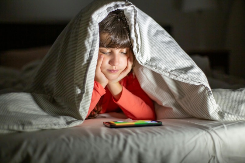 Teenager lying under a blanket propped up on their hands, looking down at a phone in a dark bedroom.