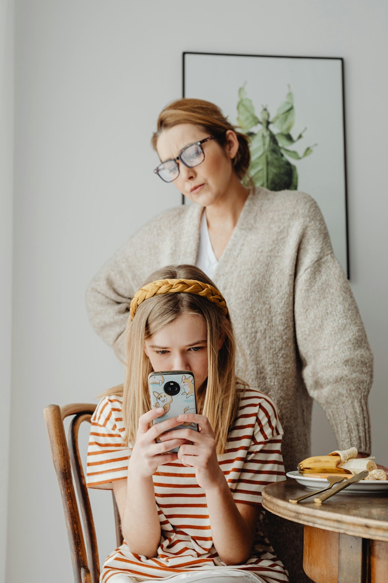 Mother standing behind her daughter, looking over her shoulder at the phone she is holding, at the breakfast table.
