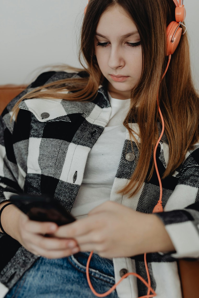Teenage girl wearing orange headphones and a checked shirt, looking down at her phone with a serious expression.
