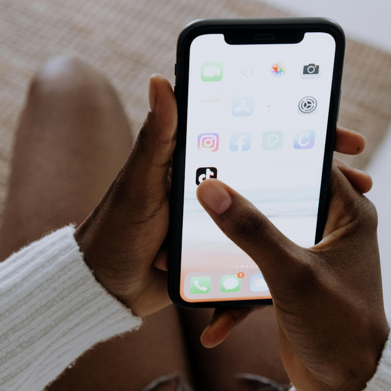 Hand holding a phone and tapping the TikTok app icon on the home screen, among other social media apps.