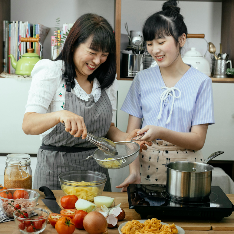 Mother and daughter smiling and cooking together in a kitchen, with vegetables and pasta on the counter.