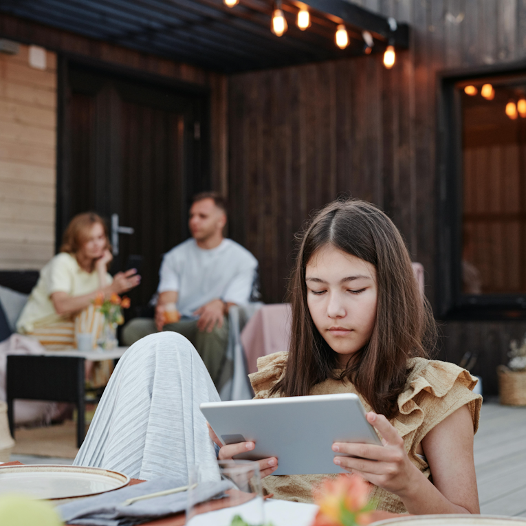 Teenage girl looking down at a tablet while sitting apart from adults chatting in the background at an outdoor evening gathering.