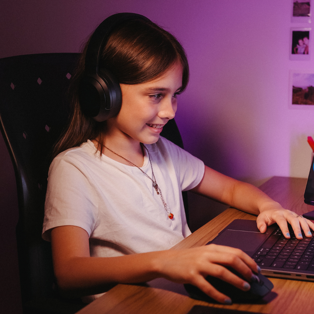 Girl wearing headphones smiling while using a laptop and mouse at a desk, lit by purple ambient lighting.