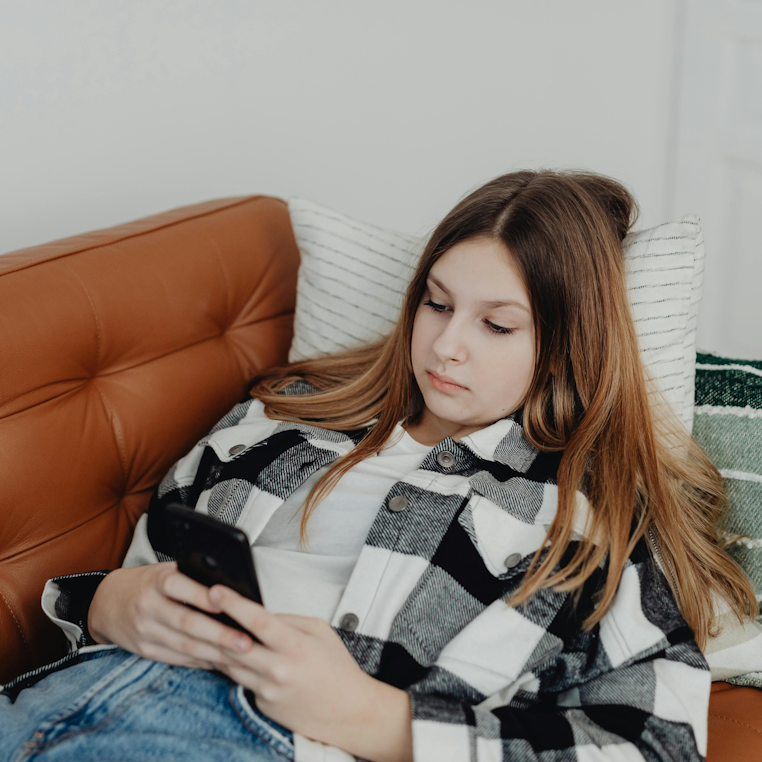 Teenage girl lying on a leather sofa, looking at her phone with a flat expression.