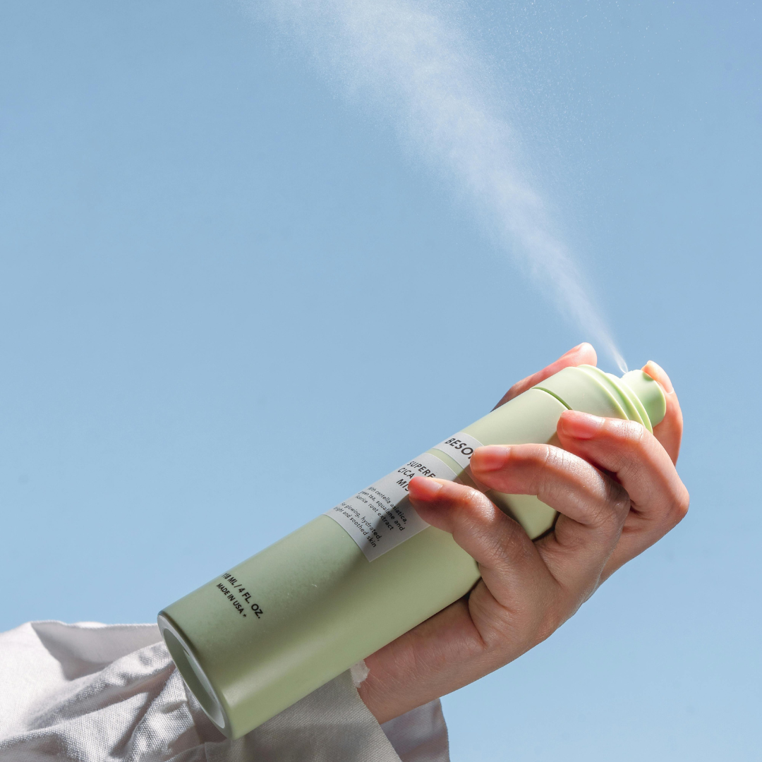 Hand holding a green spray can, spraying it into the air against a blue sky background.