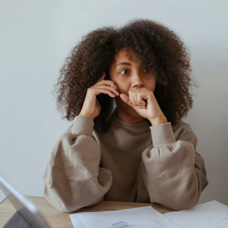 Woman with curly hair sitting at a desk, biting her fist while on a phone call, with a worried expression.