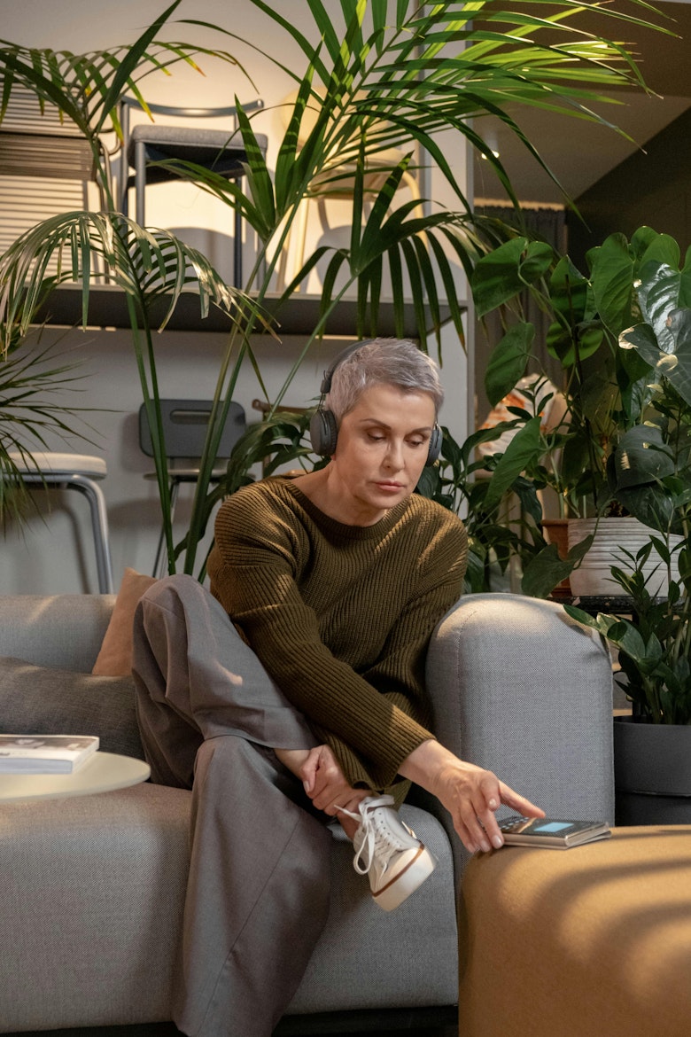 Woman with grey hair wearing headphones, sitting on a sofa surrounded by plants, selecting something on her phone.