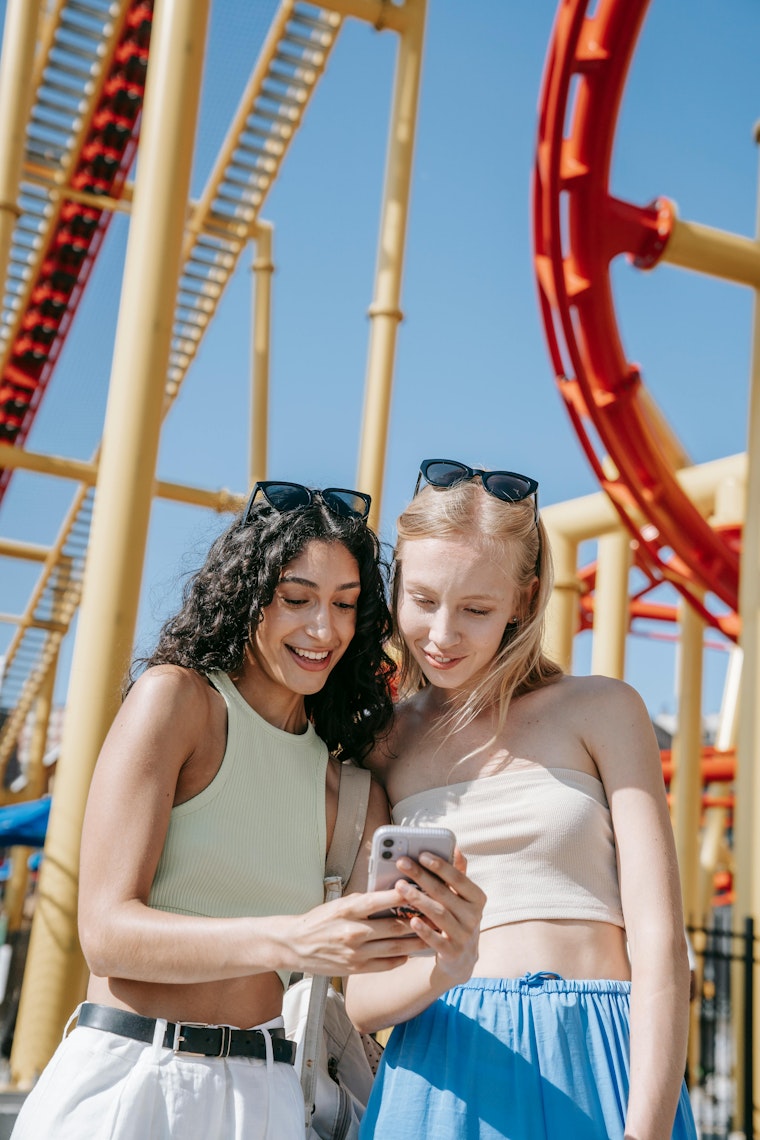 Two friends laughing while looking at a phone together, standing in front of a rollercoaster at a theme park.