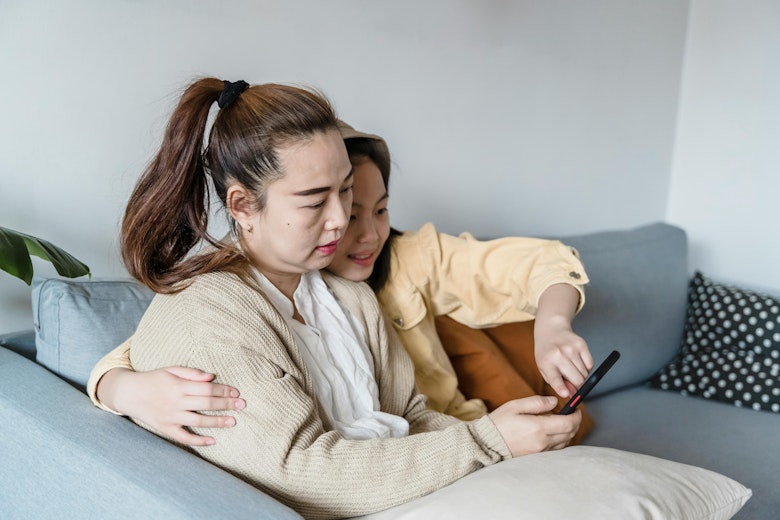 Mother with her arm around her daughter, both sitting on a sofa looking at a phone together.