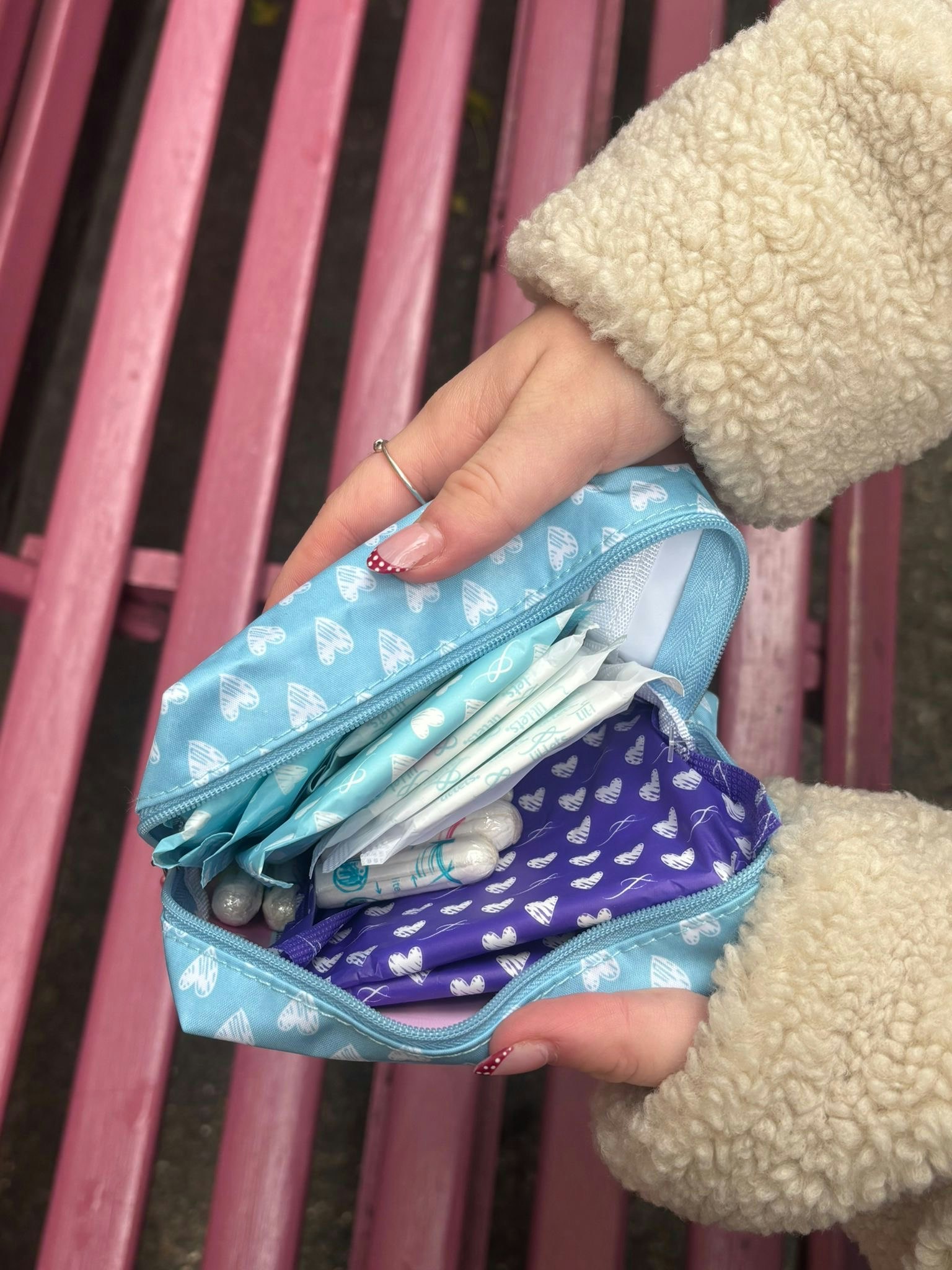 Hands opening a blue heart-patterned pouch filled with period pads and tampons, on a pink bench outdoors.