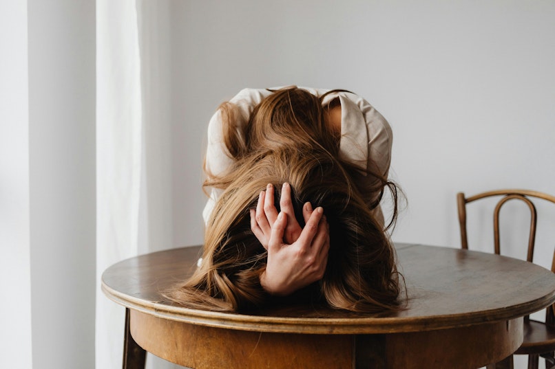 Teenager with long hair resting her head face-down on a wooden table, looking stressed.