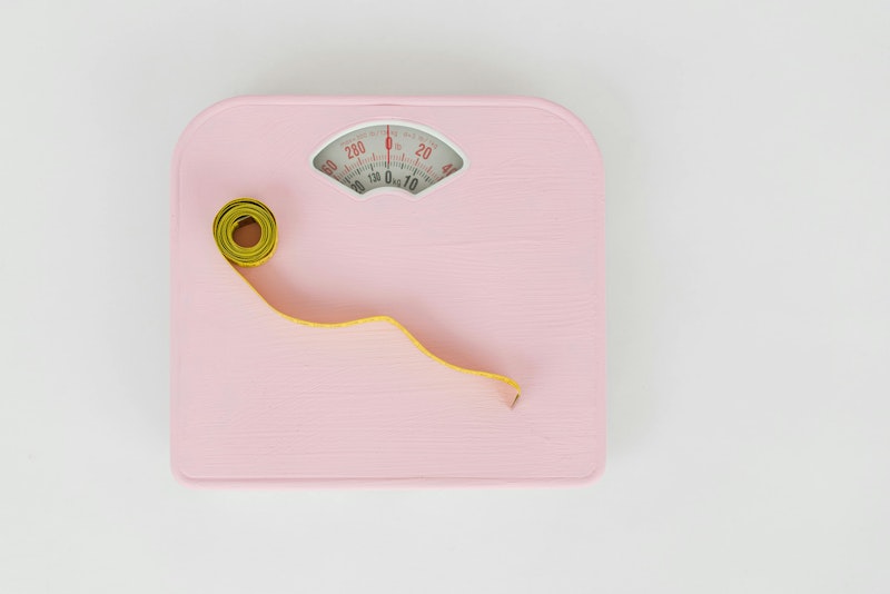 Pink bathroom scale with a yellow tape measure coiled on top of it, on a plain background.