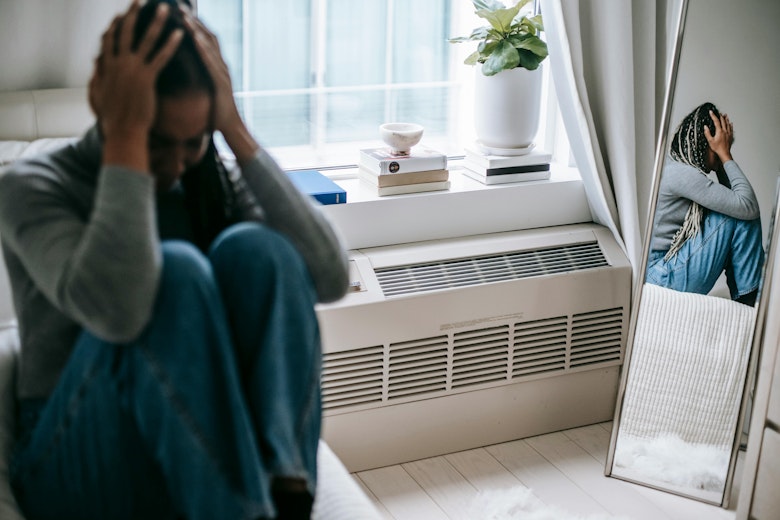 Teenage girl sitting with her head in her hands, looking distressed, by a window with a mirror beside her.