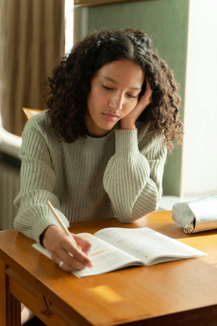 Teenage girl with curly hair sitting at a wooden desk, writing in a notebook with a pencil.