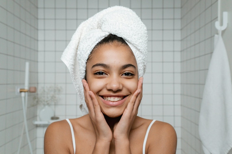 Teenage girl with a towel wrapped around her hair, smiling with her hands on her cheeks in a bathroom.