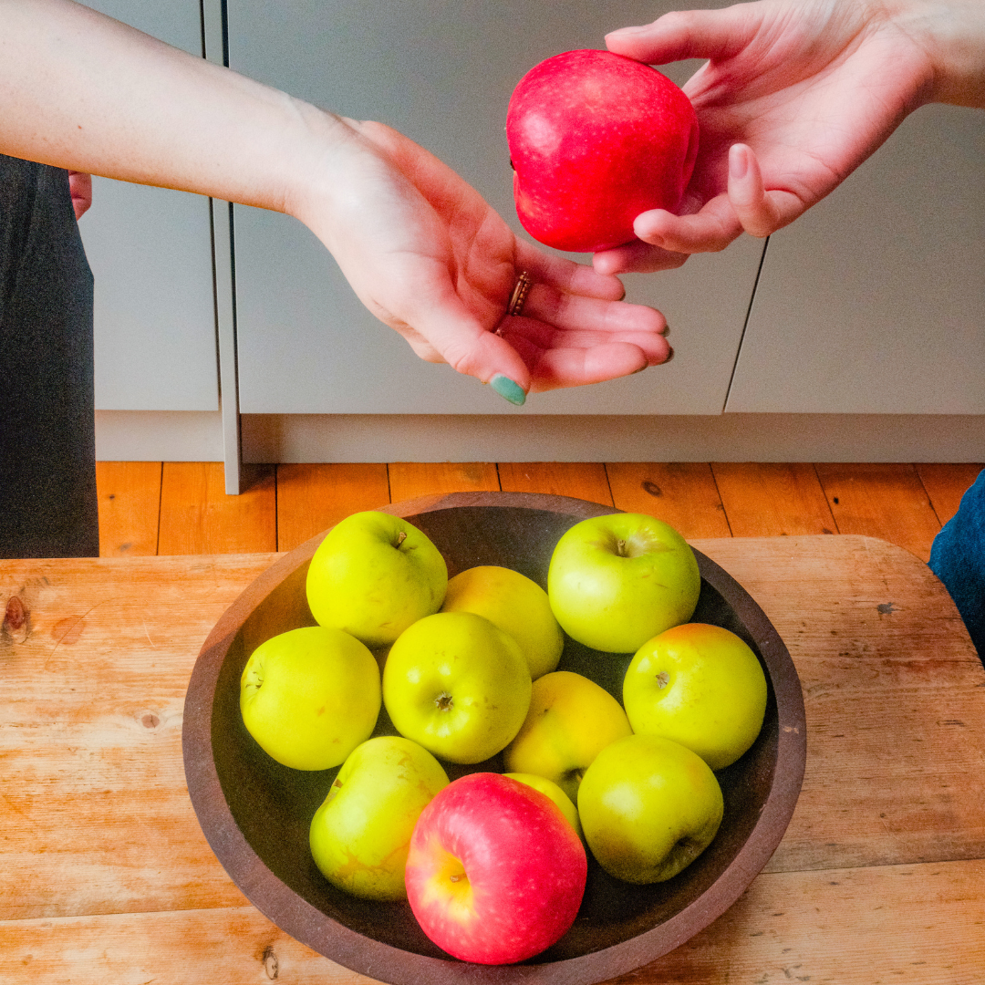 Two hands passing a red apple over a wooden bowl filled with green and red apples on a table.