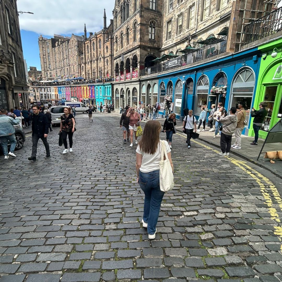 Teenager walking alone along a cobbled street lined with colourful buildings and shops, with other pedestrians nearby.
