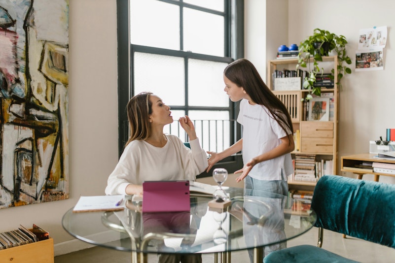 Mother sitting at a table gesturing while her daughter stands beside her talking, both with tense expressions.