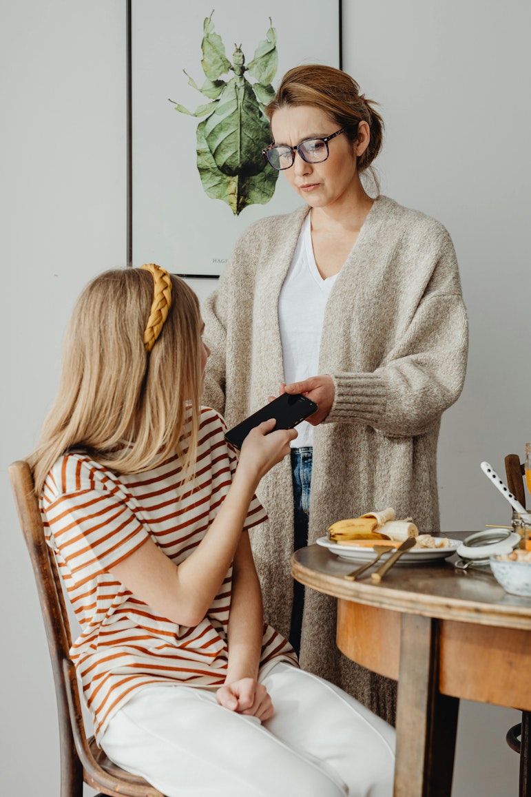 Mother standing and taking a phone from her daughter, who is sitting at a breakfast table.