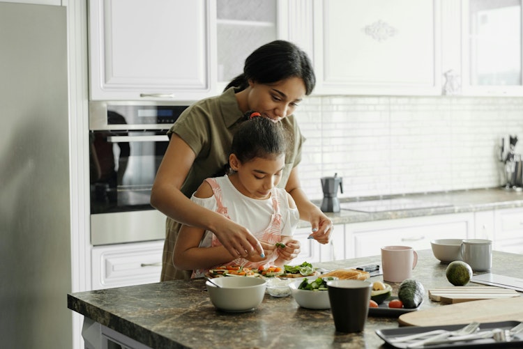 Mother helping her daughter prepare food together at a kitchen counter filled with vegetables and bowls.