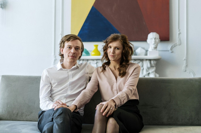A man and woman sitting together on a sofa, both looking at the camera, in front of a colourful abstract painting.