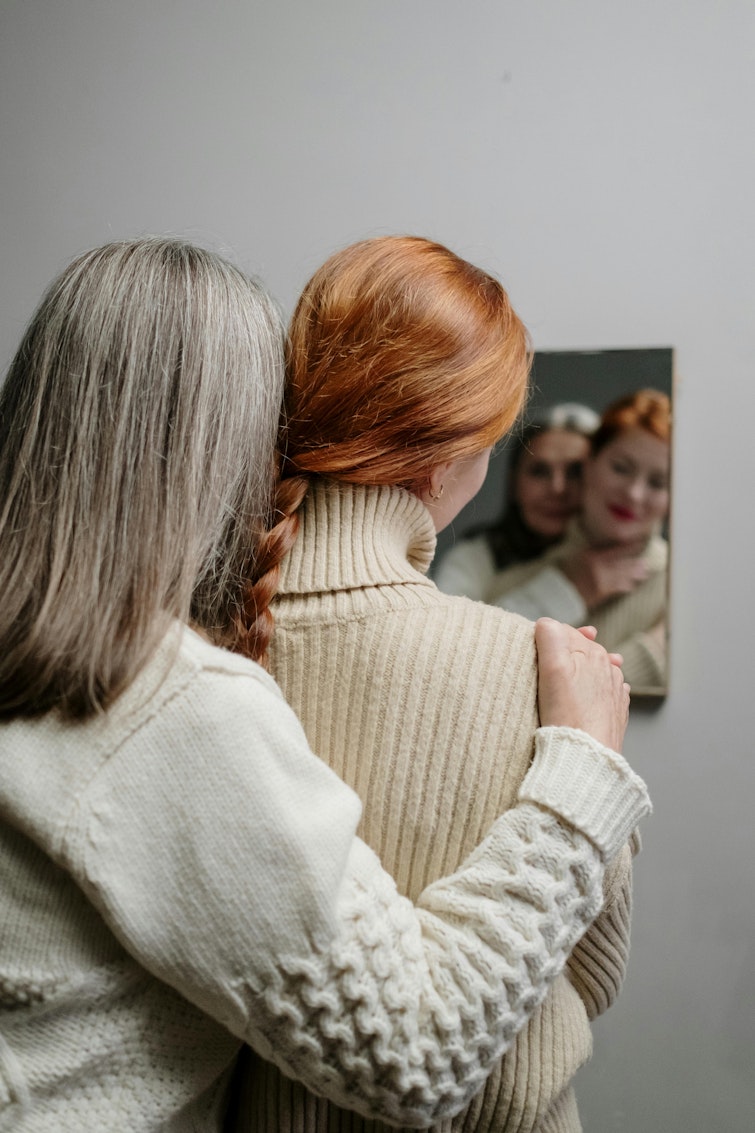 Mother with grey hair hugging her daughter, who has red hair, both reflected in a mirror.