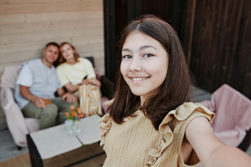 Teenage girl taking a selfie and smiling, with her parents sitting together on a sofa in the background.