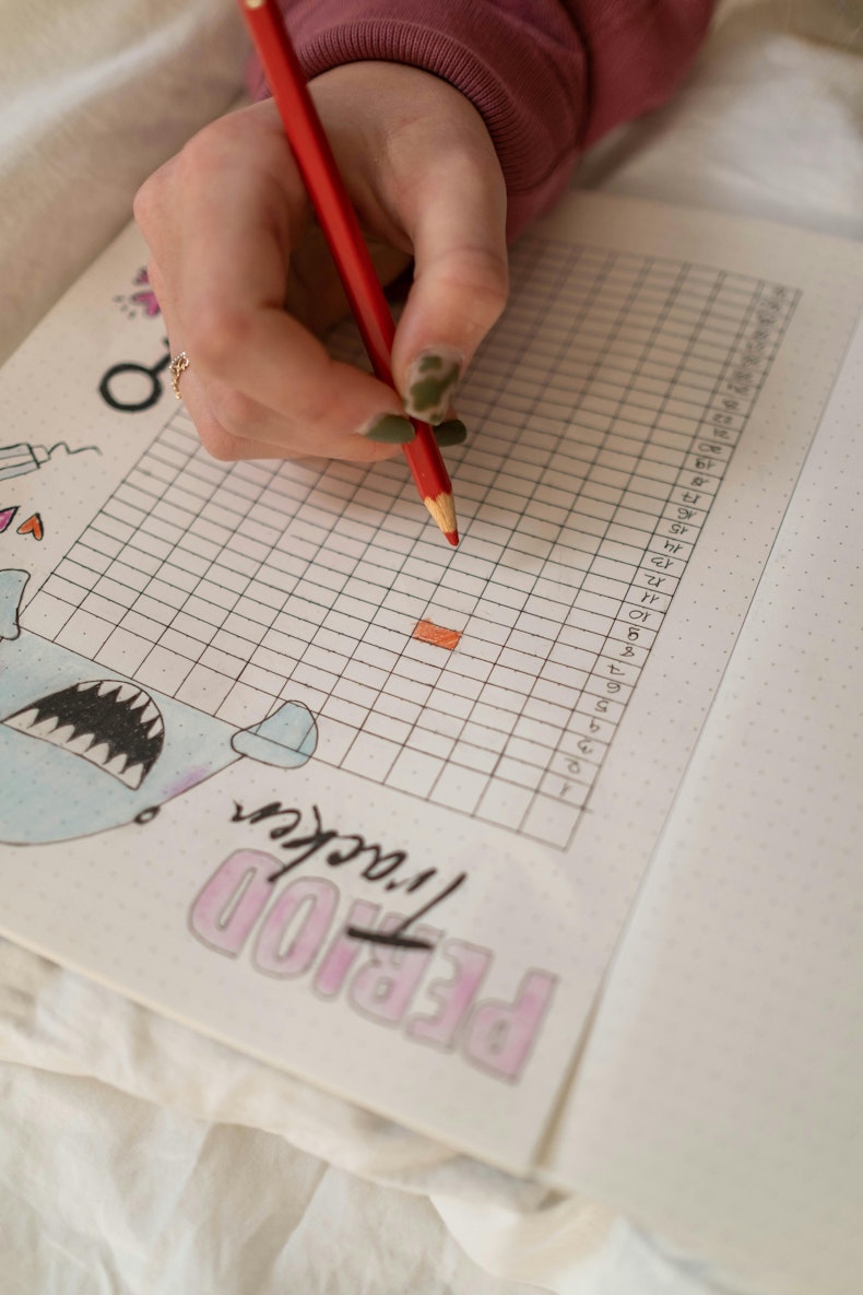 Hand holding a red pencil filling in a paper period tracker grid, with a decorated "period tracker" cover visible.