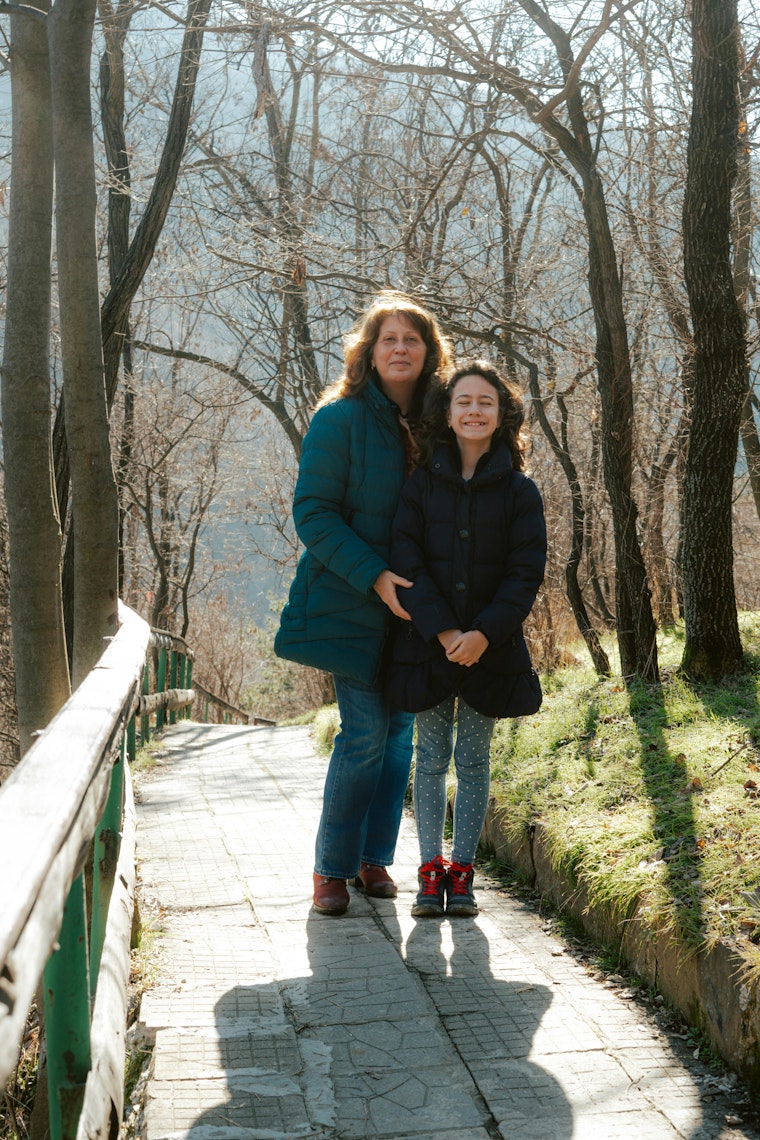 Mother and daughter smiling together on a path lined with bare trees.