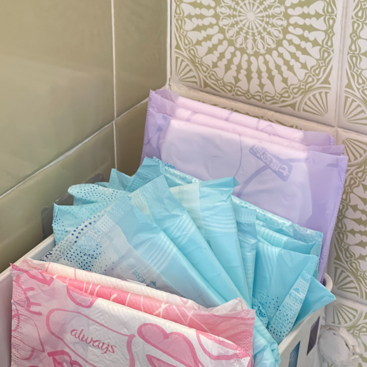 Various packets of period pads stored in a basket against a patterned tiled wall.