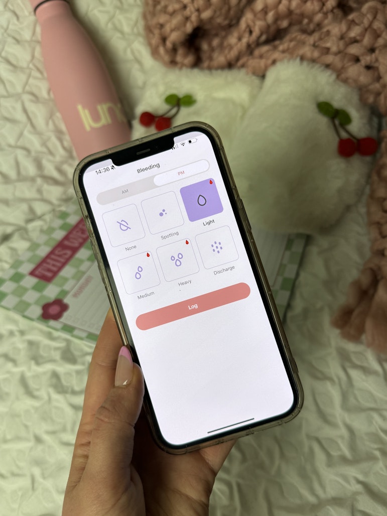 Hand holding a phone showing the luna app's bleeding tracker screen, with a pink water bottle in the background.