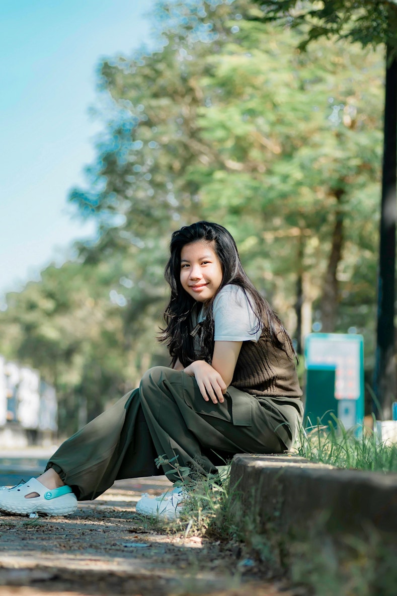 Teenage girl sitting on a low wall smiling at the camera, wearing a green outfit with trees in the background.