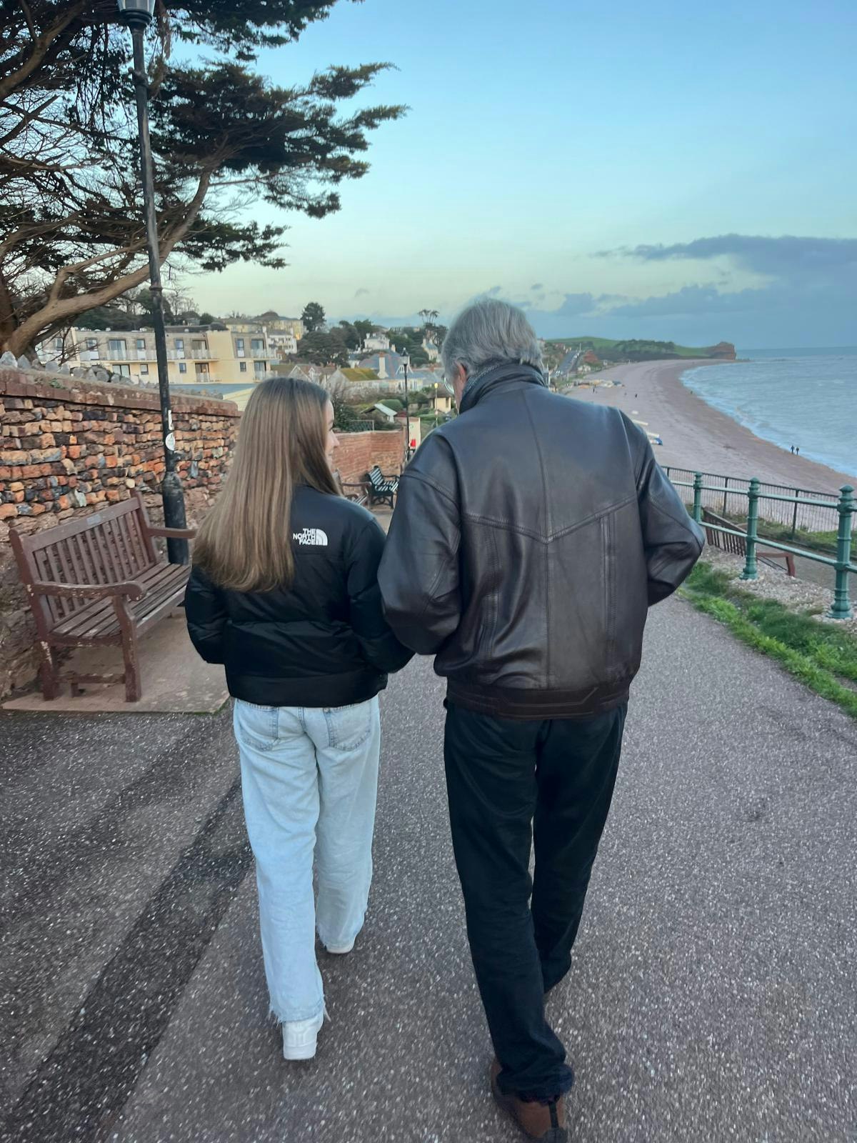Father and teenage daughter walking along a seafront path, seen from behind.