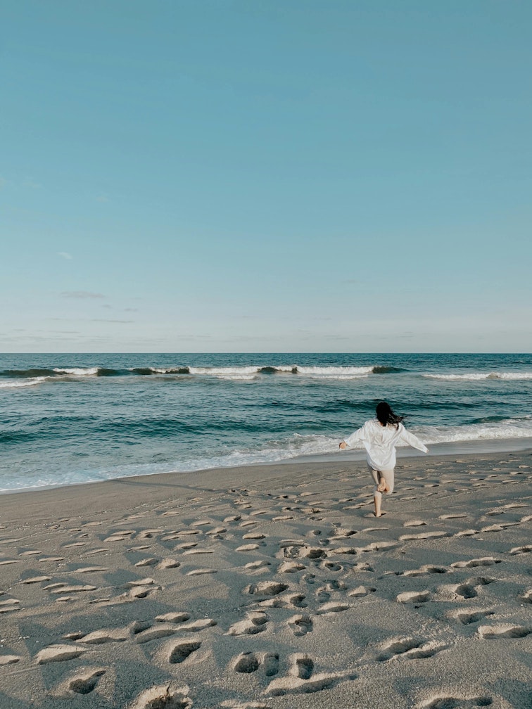 Girl in a white cover-up running along the shoreline of a beach towards the sea.