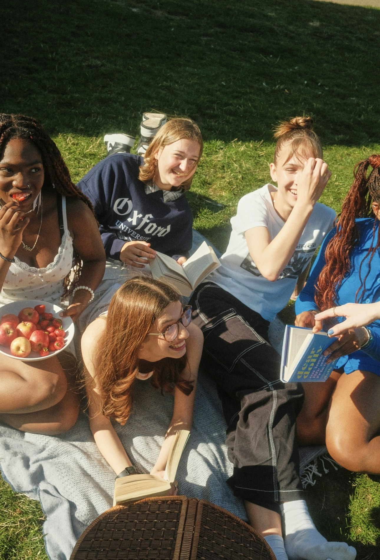 Group of teenage girls sitting on a picnic blanket in a park, laughing and reading books together with a bowl of fruit nearby.