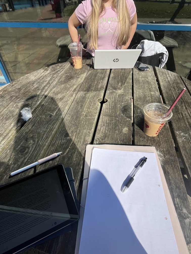 Teenage girl sitting at an outdoor picnic table with a laptop, notebook, tablet and iced drinks, studying in the sun.